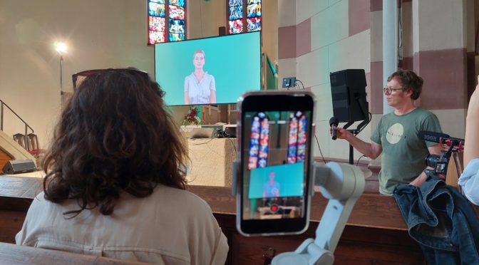 Eine KI auf einer Leinwand leitet durch den Gottesdienst, beobachtet von Menschen, Kameras und Mikrofonen.